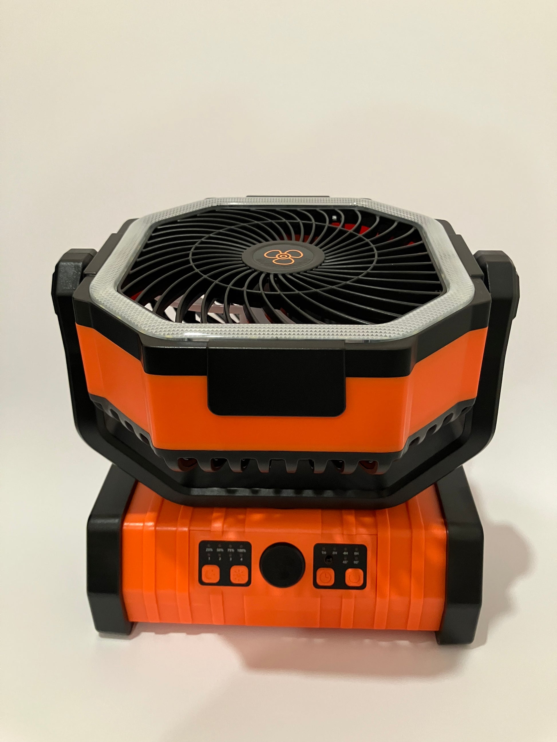 A rugged portable fan with a black and orange design, featuring a front-facing fan grille and a side battery/power unit with control buttons, shown against a plain off-white background.